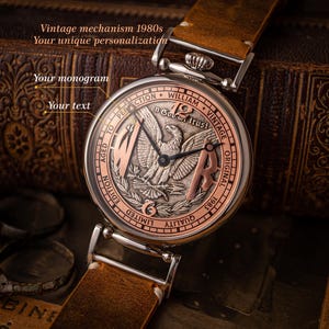May include: A vintage pocket watch with a brown leather strap. The watch face features a silver eagle with the words "In God We Trust" and "1983 Original Vintage".