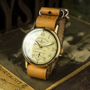 May include: A gold-tone wristwatch with a tan leather band. The watch face has black Roman numeral hour markers and a white background. The watch face also features the text "УРМАНСКИЕ" and "12 АПРЕЛЯ 1961 ЮРИЙ ГАГАРИН".