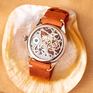 K&ouml;nnte beinhalten: Eine silberne Skelettuhr mit einem braunen Lederband. Das Zifferblatt der Uhr ist offen und zeigt das komplizierte Uhrwerk.