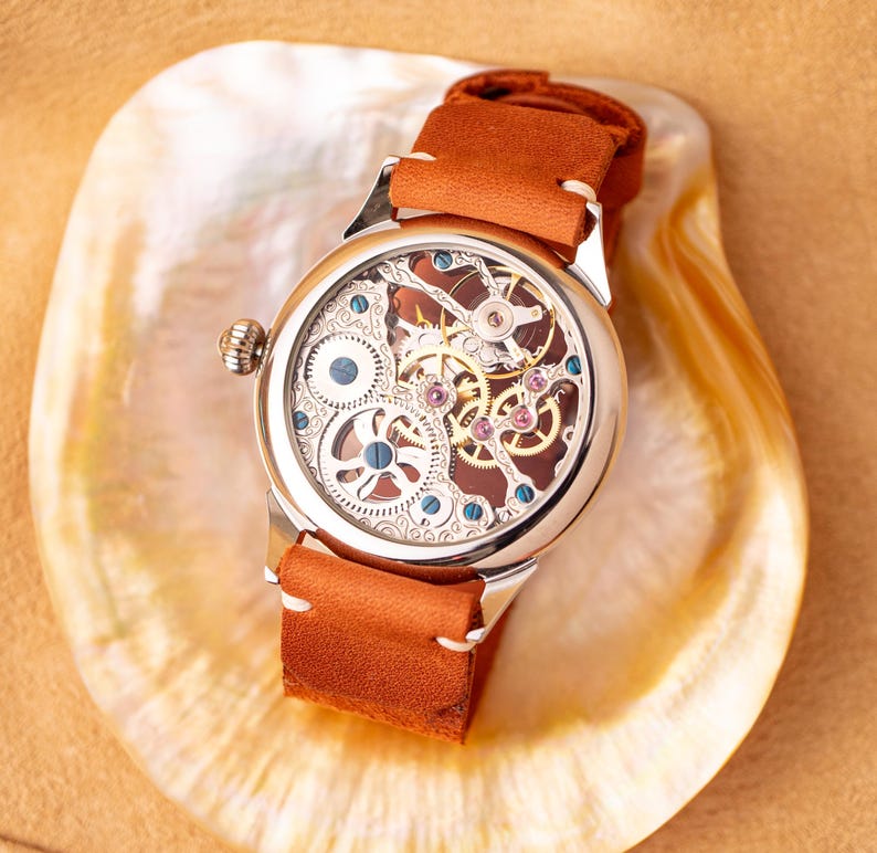 K&ouml;nnte beinhalten: Eine silberne Skelettuhr mit einem braunen Lederband. Das Zifferblatt der Uhr ist offen und zeigt das komplizierte Uhrwerk.