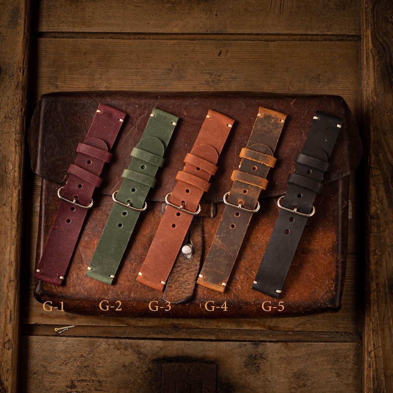 May include: Five leather watch straps in different colors, including burgundy, green, brown, dark brown, and black. The straps are laid out on a brown leather surface. The straps are labeled G-1 through G-5.