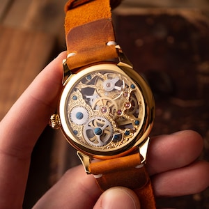 Op de afbeelding: Een goudkleurig skeleton horloge met een bruine leren band. De wijzerplaat toont ingewikkelde mechanische componenten, met blauwe en rode accenten. De band is warm bruin van kleur en het horloge wordt in een hand gehouden.