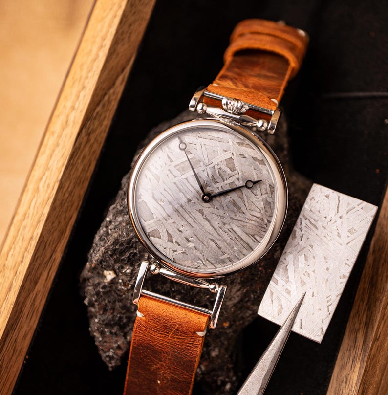 May include: A silver wristwatch with a brown leather band. The watch face is made of a meteorite with a unique pattern. The watch is displayed in a wooden box with a piece of meteorite next to it.
