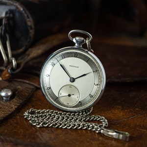 Può includere: Un orologio da tasca argentato con quadrante bianco e lancette nere. L'orologio ha una catena e le parole "МОЛНИЯ" stampate sul quadrante.