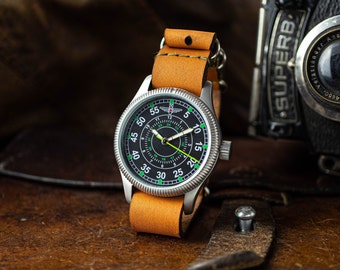 Könnte beinhalten: Eine silberne Armbanduhr mit schwarzem Zifferblatt und grünen Markierungen. Die Uhr hat ein braunes Lederband und eine silberne Schließe. Die Uhr liegt auf einer braunen Lederoberfläche.