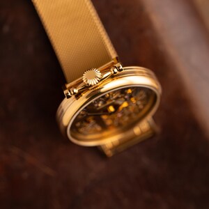 May include: A gold-toned wristwatch with a mesh band. The watch face is visible, showcasing intricate internal mechanisms. The watch's crown and case are also gold, creating a cohesive and luxurious aesthetic. The watch is set against a dark brown background.