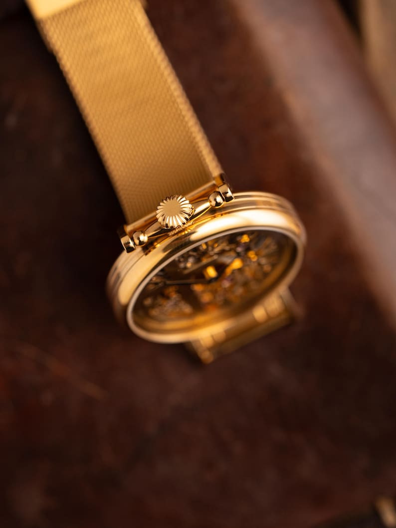 May include: A gold-toned wristwatch with a mesh band. The watch face is visible, showcasing intricate internal mechanisms. The watch's crown and case are also gold, creating a cohesive and luxurious aesthetic. The watch is set against a dark brown background.