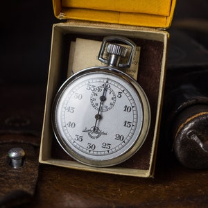 May include: A silver stopwatch with a white face and black markings. The stopwatch is in a yellow box with a white interior. The stopwatch has a black hand and a black second hand.