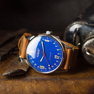 Puede incluir: Un reloj de pulsera plateado con una esfera azul y números blancos. El reloj tiene una correa de cuero marrón y las palabras "Raketa" y "Cul 2023 H" están impresas en la esfera.