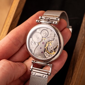 May include: A silver pocket watch with a mesh metal band. The watch face is open, revealing the intricate inner workings of the watch.