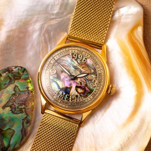 May include: A gold-tone wristwatch with a mother-of-pearl face. The face has the words "Those who believe in their dreams, the future belongs to them" and the year "1996" printed on it.