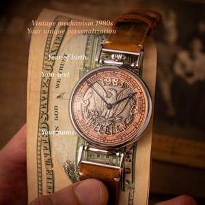 May include: A silver pocket watch with a brown leather band. The watch face features a silver eagle with the words "Dreams The Future Belongs To Those Who Believe In Their" and "1983" around the eagle. The watch is on top of a dollar bill.