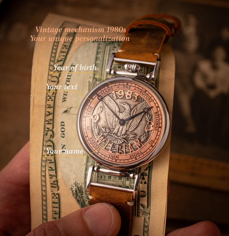 May include: A silver pocket watch with a brown leather band. The watch face features a silver eagle with the words "Dreams The Future Belongs To Those Who Believe In Their" and "1983" around the eagle. The watch is on top of a dollar bill.