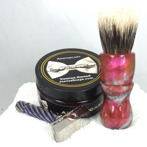 May include: A shaving set with a silver razor, a blue and silver handle razor, a red and silver shaving brush with a white brush head, and a black jar of shaving cream with a white bow tie and the text "Apothecary Jeeves of Hudson Street LLC Veteran Owned JeevesSoaps.com"