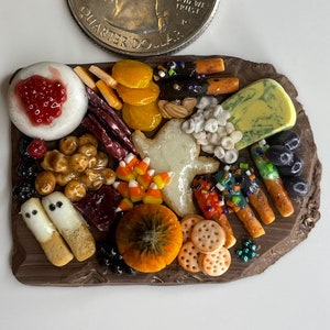 Op de afbeelding: Een miniatuur Halloween-charcuteriebord met een verscheidenheid aan snacks, waaronder kaas, crackers, fruit, candy corn en koekjes in de vorm van een spook. Het bord is gemaakt van bruine klei en is ongeveer zo groot als een kwartje.