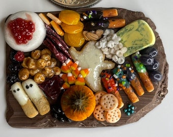 May include: A miniature Halloween-themed charcuterie board with a variety of snacks, including cheese, crackers, fruit, candy corn, and ghost-shaped cookies. The board is made of brown clay and is about the size of a quarter.
