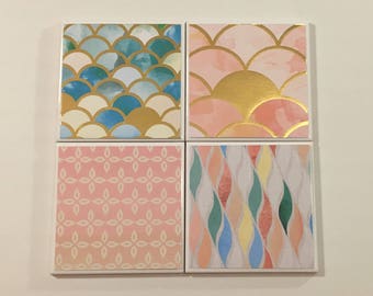 May include: Set of four square coasters with different geometric patterns. The coasters feature a pink, blue, and gold color scheme.
