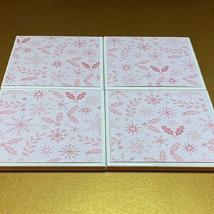 Peut inclure: Ensemble de quatre sous-verres en c&eacute;ramique blanche avec un motif de flocons de neige et de feuilles de houx rose et blanc.