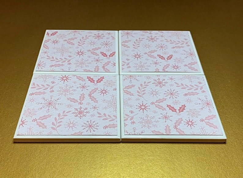 Peut inclure: Ensemble de quatre sous-verres en c&eacute;ramique blanche avec un motif de flocons de neige et de feuilles de houx rose et blanc.
