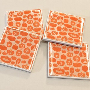 May include: Four square coasters with a glossy finish, each featuring a repeating pattern of orange pumpkins, bats, spiders, and witch hats on a light peach background. The letter D is printed in black in the upper right corner of the image.