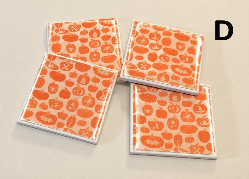 May include: Four square coasters with a glossy finish, each featuring a repeating pattern of orange pumpkins, bats, spiders, and witch hats on a light peach background. The letter D is printed in black in the upper right corner of the image.