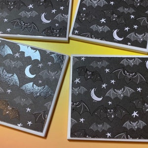 May include: Four square coasters featuring a repeating pattern of bats, crescent moons, and stars on a dark grey background. The coasters have a white border and a smooth surface, with some bats rendered in a metallic silver or gold finish.