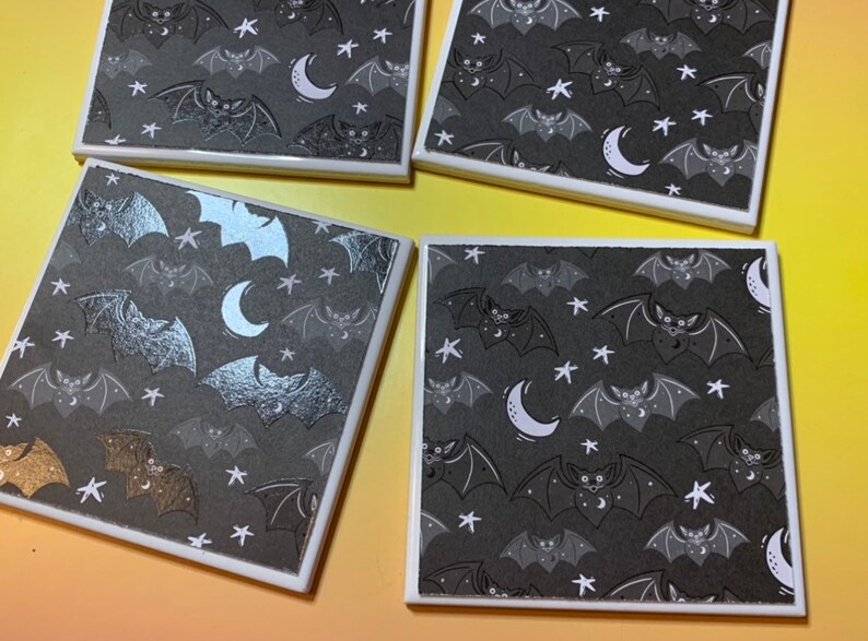 May include: Four square coasters featuring a repeating pattern of bats, crescent moons, and stars on a dark grey background. The coasters have a white border and a smooth surface, with some bats rendered in a metallic silver or gold finish.