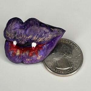 K&ouml;nnte beinhalten: Lila und goldene Glitzerlippen mit wei&szlig;en Z&auml;hnen und rotem, tropfendem Blut. Die Lippen sind etwa so gro&szlig; wie eine 25-Cent-M&uuml;nze.