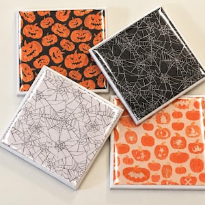 May include: A set of four square coasters with glossy finishes. Two coasters feature orange pumpkins on a black background, one has a black spiderweb pattern on a white background, and one has a white spiderweb pattern on a black background.