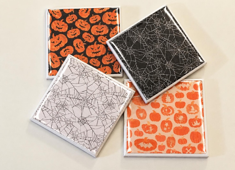 May include: A set of four square coasters with glossy finishes. Two coasters feature orange pumpkins on a black background, one has a black spiderweb pattern on a white background, and one has a white spiderweb pattern on a black background.