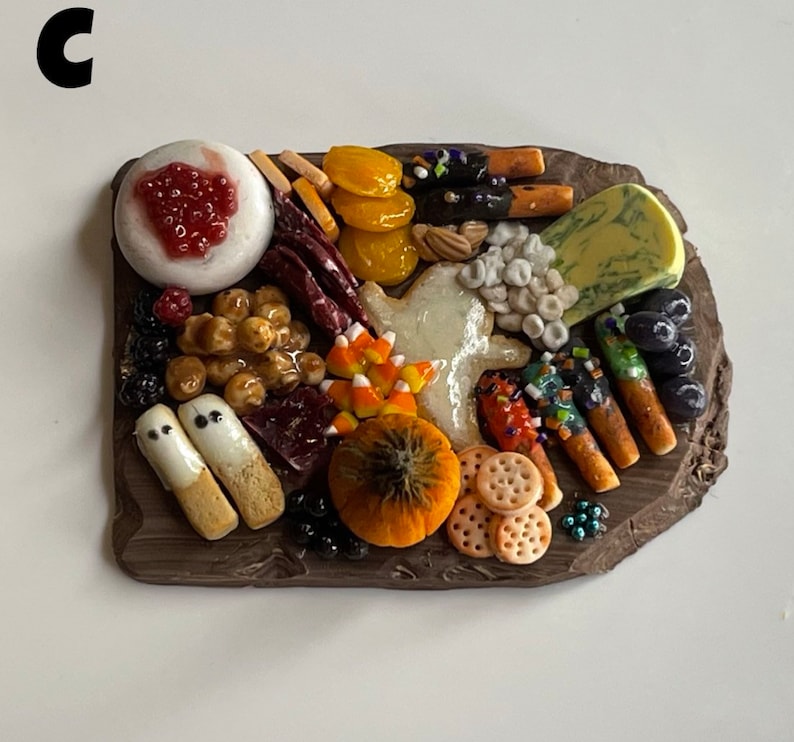 Op de afbeelding: Een miniatuur Halloween-charcuteriebord met een verscheidenheid aan etenswaren, waaronder kaas, crackers, fruit en snoep. Het bord is gemaakt van bruin klei en heeft een rustieke uitstraling.