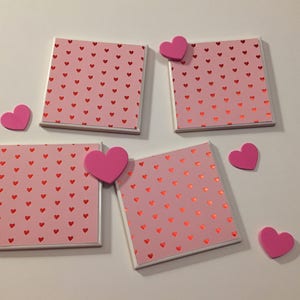 May include: Four pink and white ceramic coasters with a red heart pattern. The coasters are surrounded by pink foam hearts.