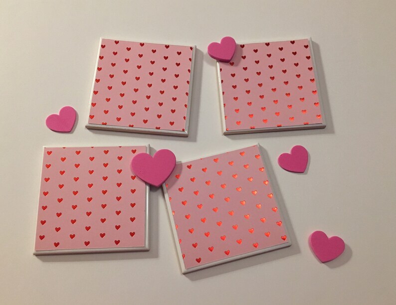May include: Four pink and white ceramic coasters with a red heart pattern. The coasters are surrounded by pink foam hearts.