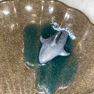 Puede incluir: Una figurita de tibur&oacute;n de color plateado en una piscina de resina de color turquesa dentro de un plato de cer&aacute;mica marr&oacute;n en forma de concha.
