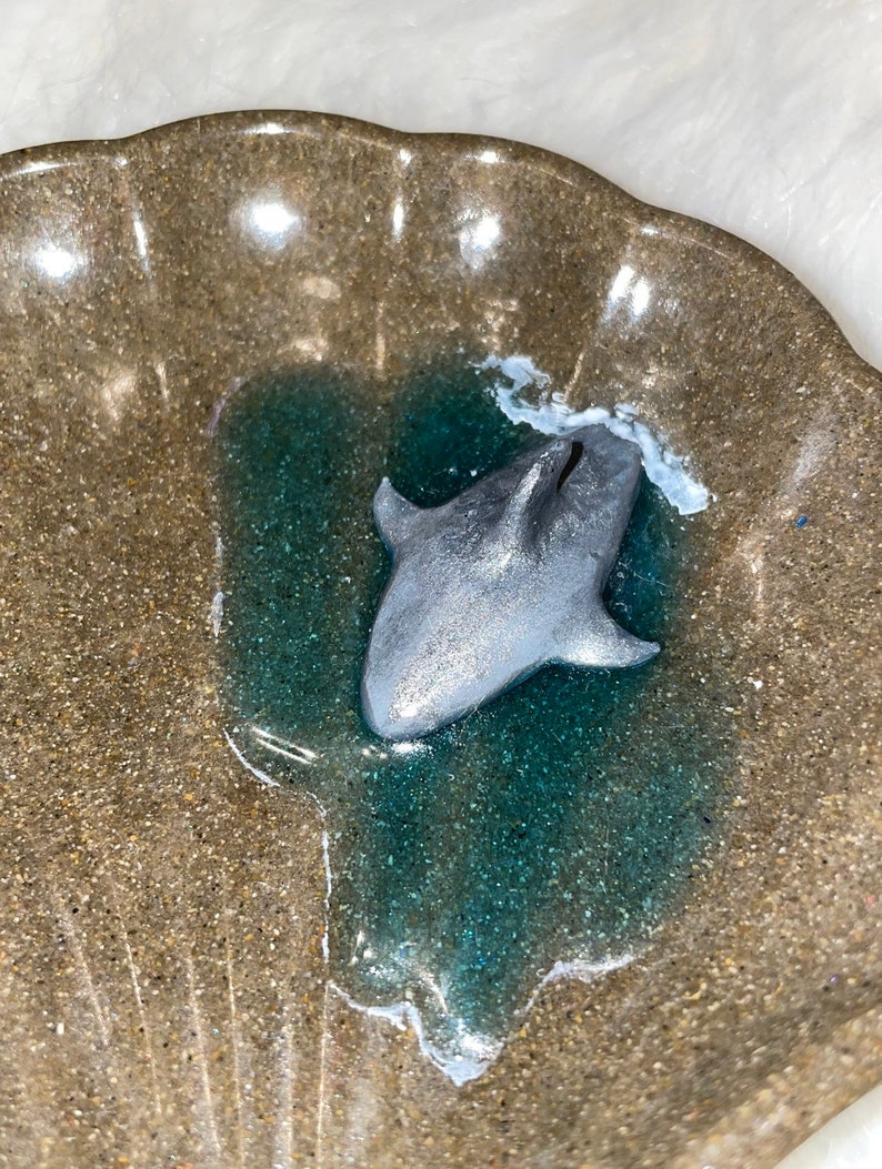Puede incluir: Una figurita de tibur&oacute;n de color plateado en una piscina de resina de color turquesa dentro de un plato de cer&aacute;mica marr&oacute;n en forma de concha.