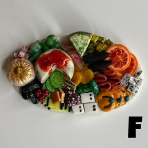 Op de afbeelding: Een miniatuur voedselplateau met een verscheidenheid aan items, waaronder een ronde kaas met rode jam, groene druiven, kiwi-schijfjes, sinaasappelschijfjes en een gouden pompoen. Het plateau bevindt zich op een wit ovaal oppervlak.