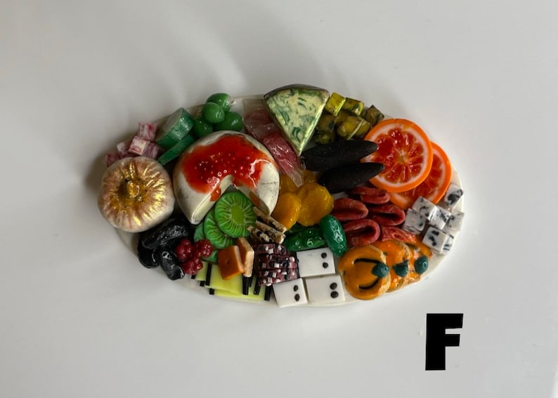 Op de afbeelding: Een miniatuur voedselplateau met een verscheidenheid aan items, waaronder een ronde kaas met rode jam, groene druiven, kiwi-schijfjes, sinaasappelschijfjes en een gouden pompoen. Het plateau bevindt zich op een wit ovaal oppervlak.