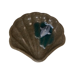 Puede incluir: Un plato de color marr&oacute;n con forma de concha de mar con una peque&ntilde;a figura de delf&iacute;n gris en su interior. El plato est&aacute; hecho de un material texturizado que se asemeja a la piedra.