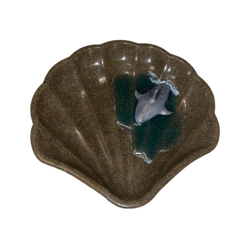 Puede incluir: Un plato de color marr&oacute;n con forma de concha de mar con una peque&ntilde;a figura de delf&iacute;n gris en su interior. El plato est&aacute; hecho de un material texturizado que se asemeja a la piedra.