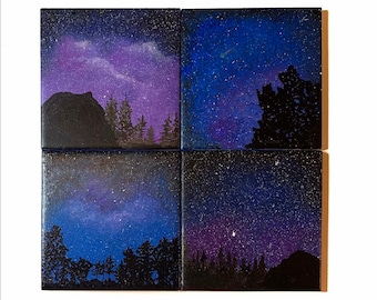 May include: Four square canvases with a night sky painted on them. Each canvas features a different view of the night sky with stars and a galaxy. The sky is painted in shades of blue, purple, and black. The canvases are arranged in a grid pattern.