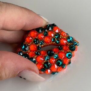 May include: A bejeweled, open-mouthed lips-shaped brooch. The brooch is covered in orange and teal-colored rhinestones, with gold-colored glitter around the edges. The brooch is held by a person's fingers.