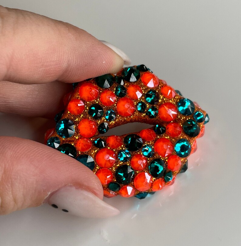 May include: A bejeweled, open-mouthed lips-shaped brooch. The brooch is covered in orange and teal-colored rhinestones, with gold-colored glitter around the edges. The brooch is held by a person's fingers.
