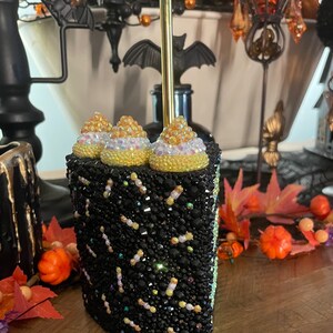May include: A black and gold cake-shaped tumbler with a gold straw. The tumbler is covered in rhinestones and has a black and gold frosting design. The tumbler is decorated with three gold and white cake toppers.