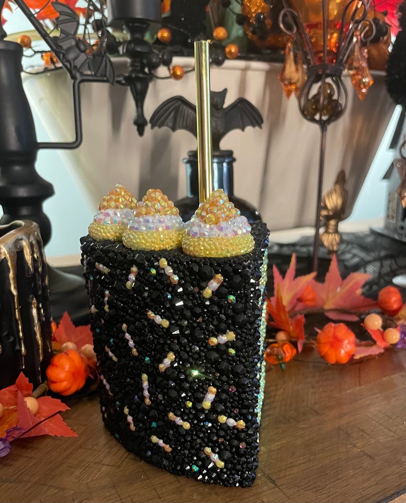 May include: A black and gold cake-shaped tumbler with a gold straw. The tumbler is covered in rhinestones and has a black and gold frosting design. The tumbler is decorated with three gold and white cake toppers.