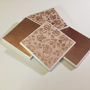 Puede incluir: Cuatro posavasos cuadrados de cer&aacute;mica con borde blanco. Dos posavasos tienen un fondo rosa con un patr&oacute;n floral dorado. Dos posavasos son de cobre macizo.