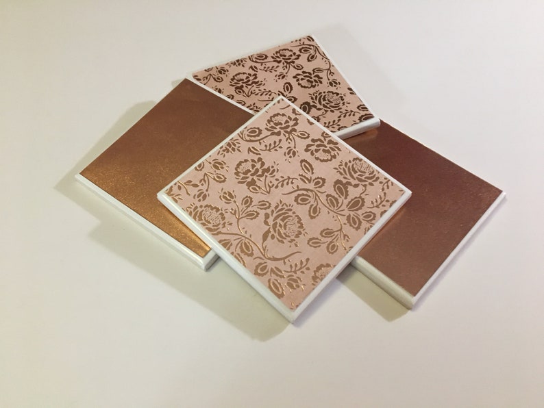 Puede incluir: Cuatro posavasos cuadrados de cer&aacute;mica con borde blanco. Dos posavasos tienen un fondo rosa con un patr&oacute;n floral dorado. Dos posavasos son de cobre macizo.