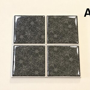 May include: Four square coasters arranged in a grid, each featuring a glossy black surface with a white spider web pattern. The letter A is positioned in the upper right corner. The coasters have a smooth, reflective finish and white edges.