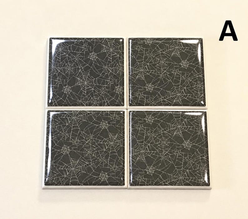May include: Four square coasters arranged in a grid, each featuring a glossy black surface with a white spider web pattern. The letter A is positioned in the upper right corner. The coasters have a smooth, reflective finish and white edges.