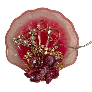 Puede incluir: Plato para joyas en forma de concha rosa con un pulpo morado, purpurina dorada y detalles de pan de oro. El plato est&aacute; lleno de joyas, incluyendo anillos, un collar y pendientes.
