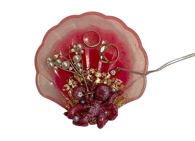 Puede incluir: Plato para joyas en forma de concha rosa con un pulpo morado, purpurina dorada y detalles de pan de oro. El plato est&aacute; lleno de joyas, incluyendo anillos, un collar y pendientes.